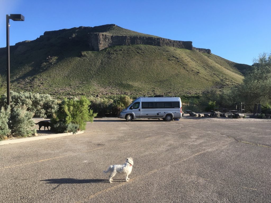 Parking lot with camper van parked in front of beautiful green hill and cliff. Also a cute Jack Russell terrier in front of the van. 