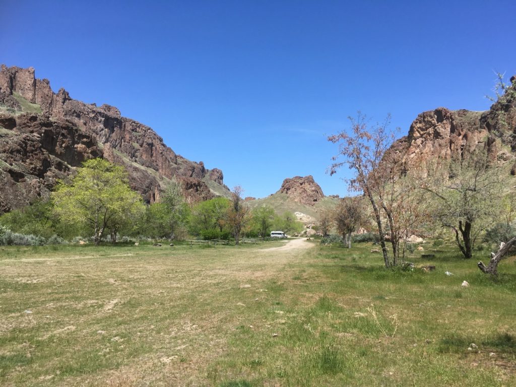 Campground view -- a long dirt road with a tiny van at the end of it, green trees, canyon walls on either side. 